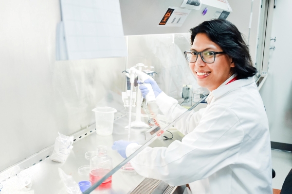 student wearing a white lab coat holds pipette equipment in his right hand and transfers a red liquid into a petri disk.