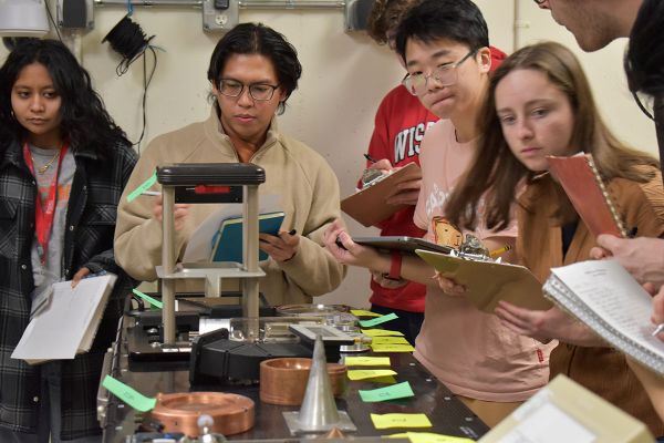 A group of students holding clipboards stand around a table taking a test. Parts of a linear accelerator are in front of them on the table.