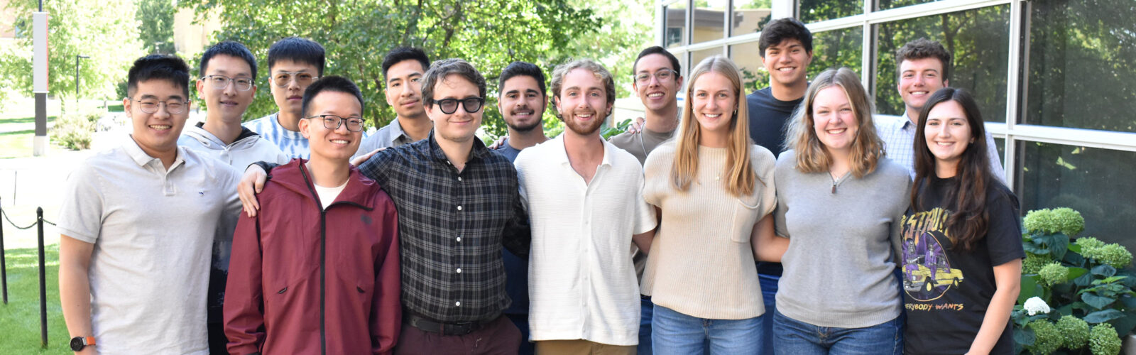 Fourteen graduate students stand outside in front of a building and gather close together.