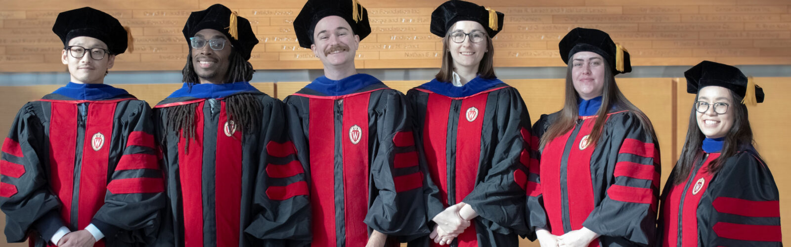 six graduate students stand together smiling wearing graduation robes that are black with red