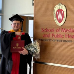 Bujar stands in graduation robes holding flowers in front of School of Medicine and Public Health sign