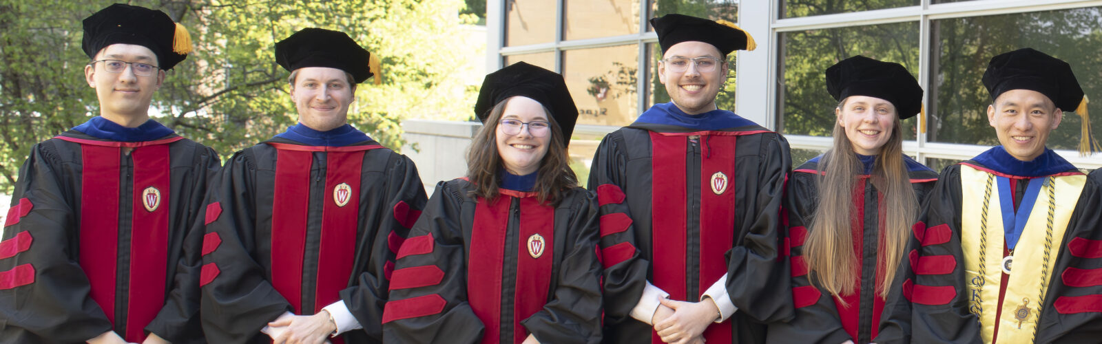 A group of people wearing red and black graduation gowns and tams stand outside on a sunny day