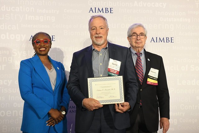 Andy stands in between two other people and he's holding an award certificate
