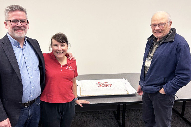 Mike, Beth and Jim stand together in front of a cake at a goodbye reception