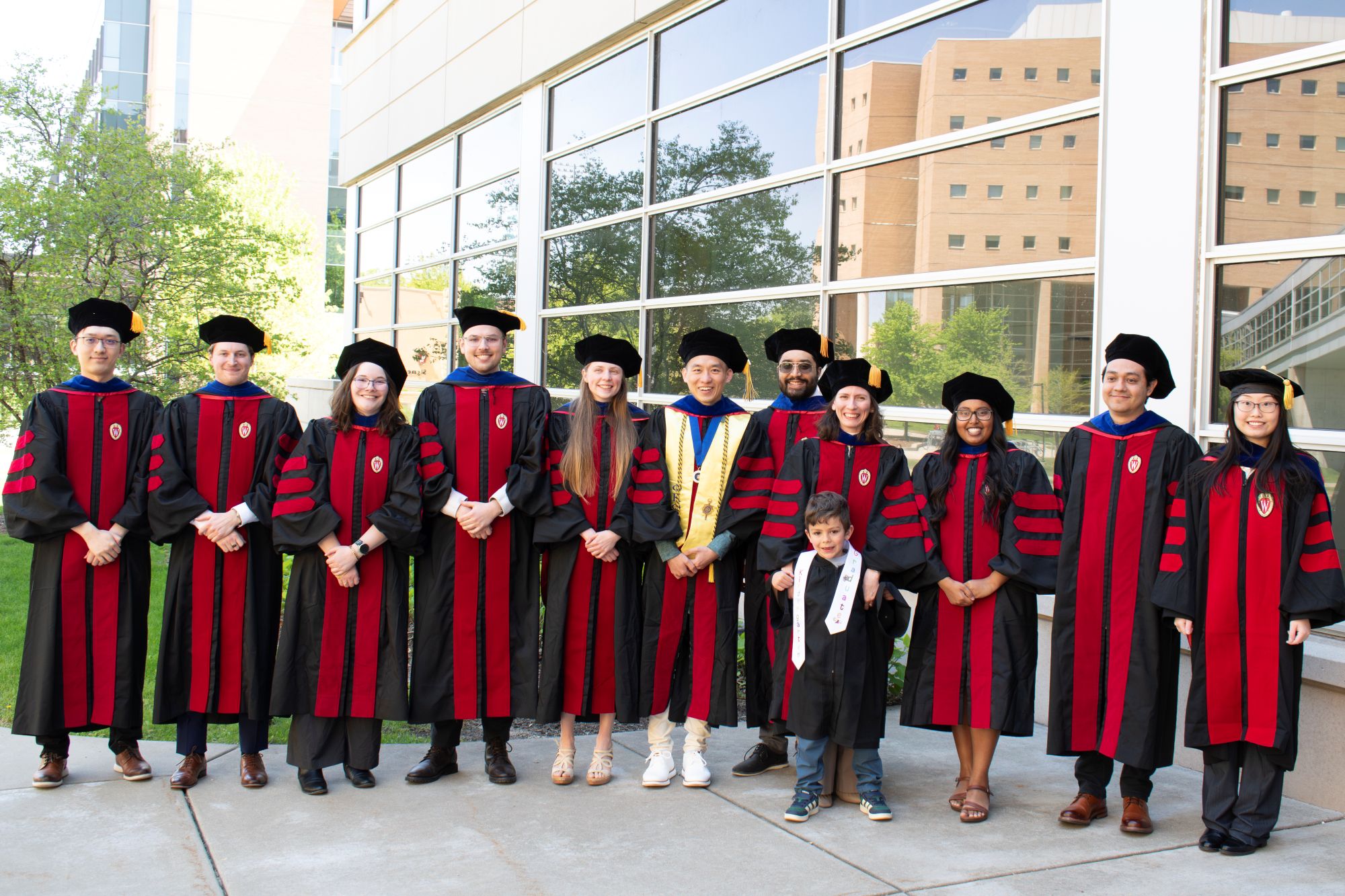 11 graduates stand together wearing black and red robes outside on a sunny day