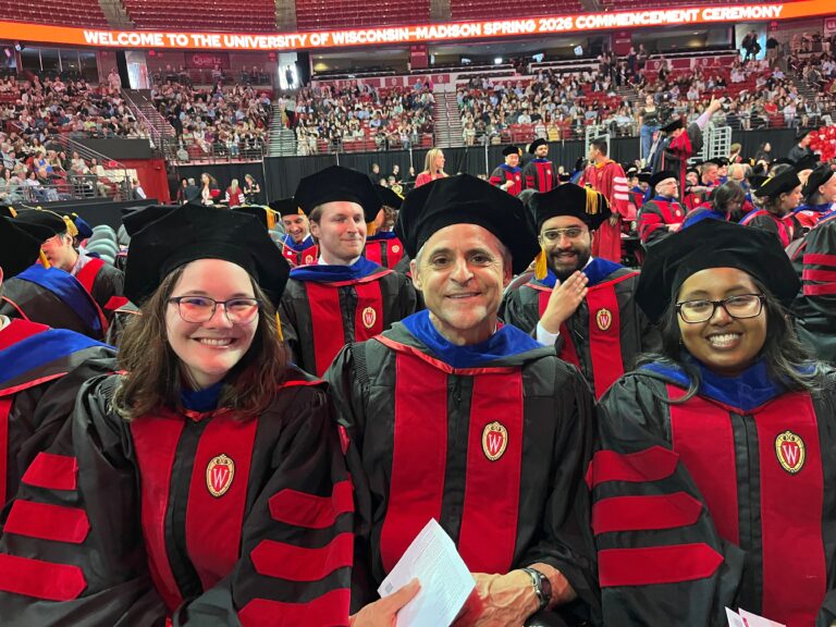 graduates and faculty wearing robes are sitting in the Kohl Center at a crowded graduation ceremony