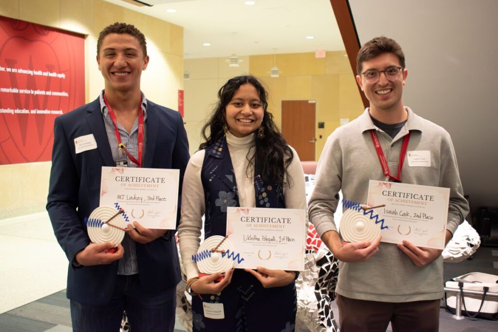 three graduate students stand in a lobby holding award certificates